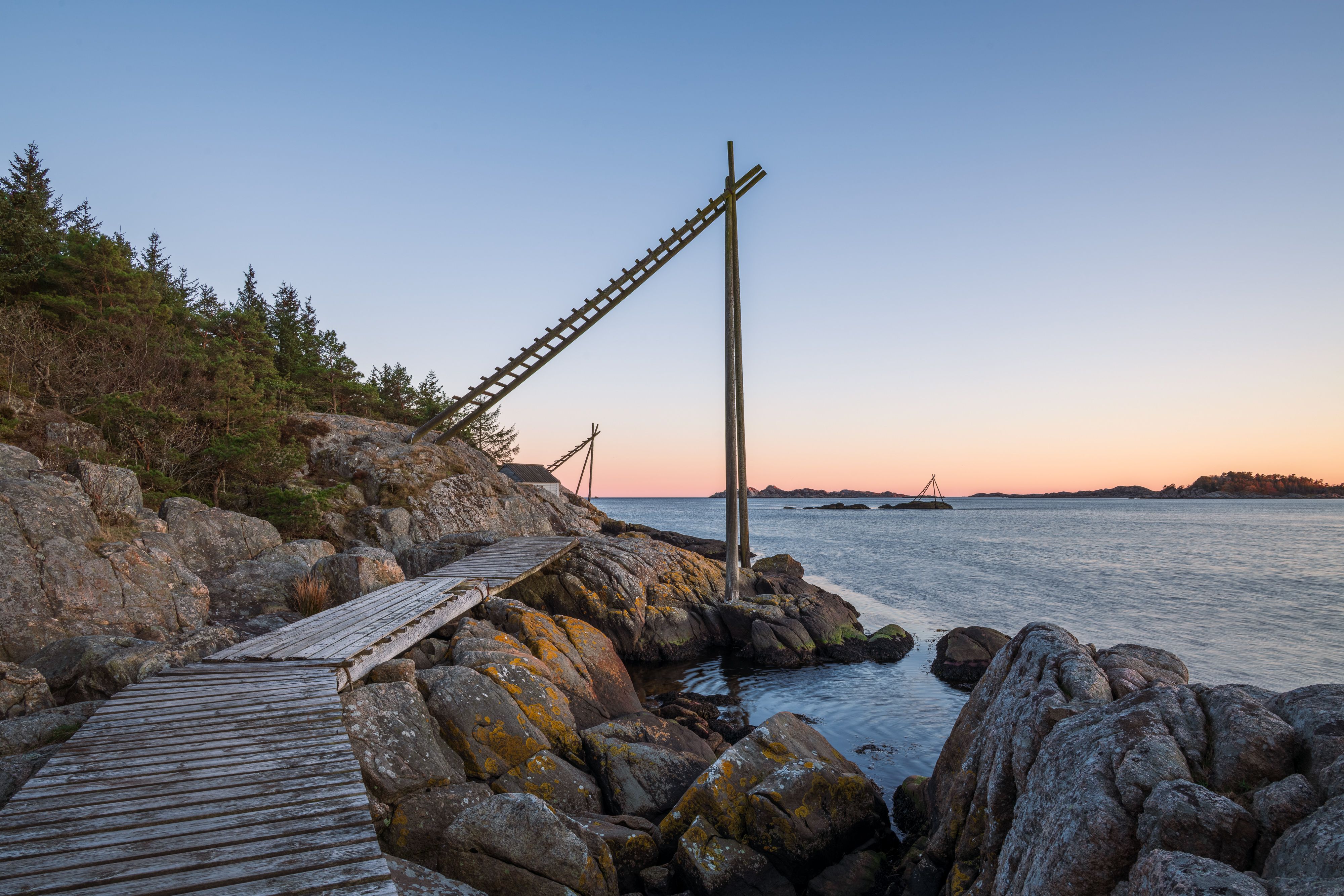 Smal trebrygge over svaberg mot rolig sjø og skjærgård i varmt kveldslys ved Mandal.