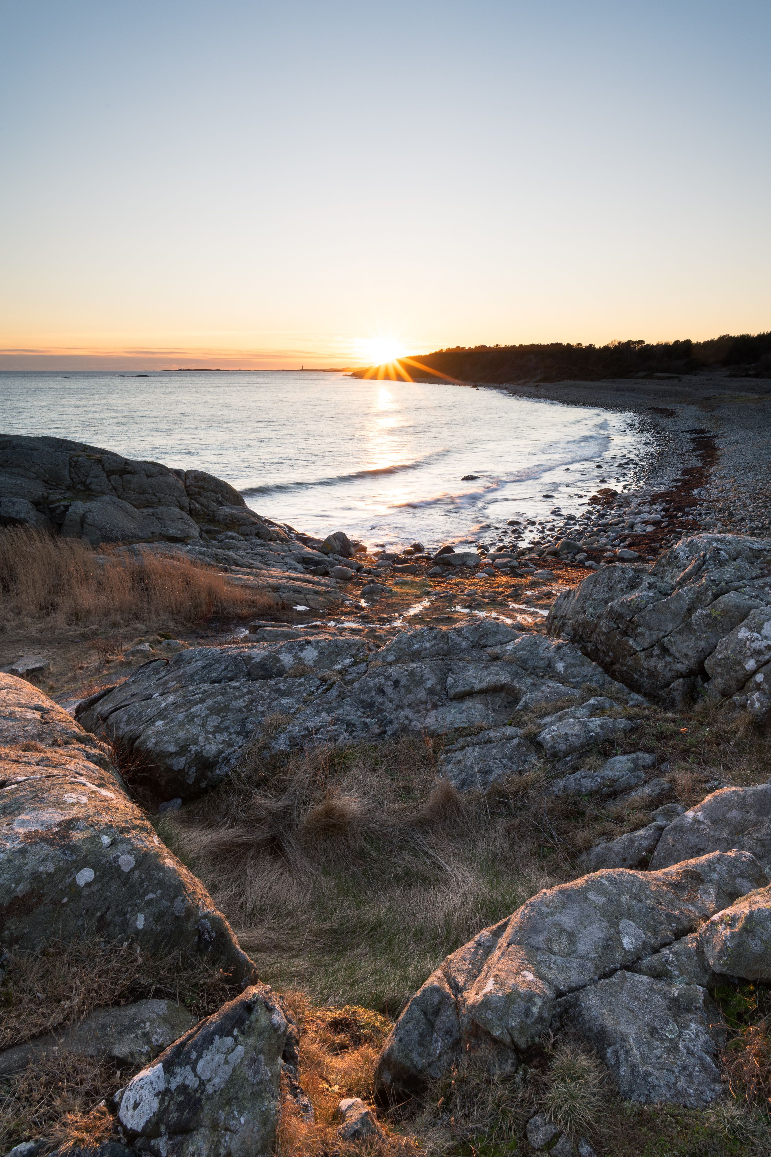 Soloppgang over en steinete strand med svaberg og vissent gress i Raet nasjonalpark.